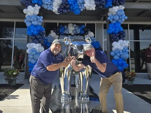 Two men holding a large chrome trophy beneath a balloon arch outside an office building