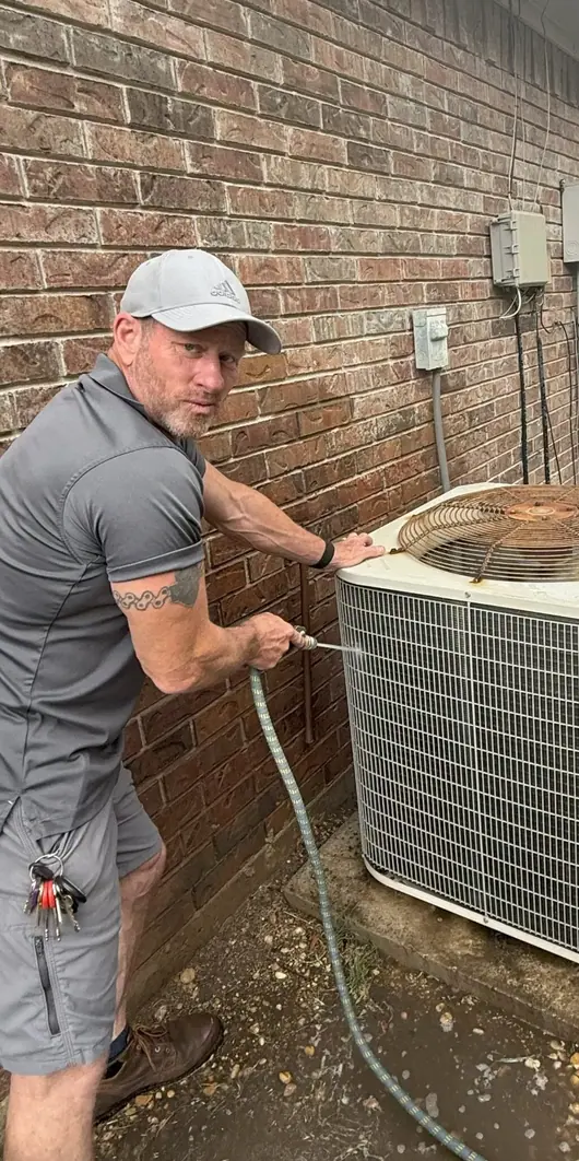 An HVAC technician rinsing the coil of an outdoor condenser unit beside a brick house