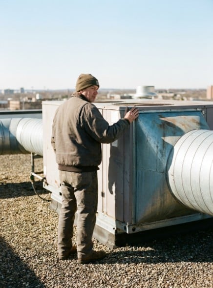 An HVAC technician on a flat roof beside a rooftop air handling unit and its duct runs