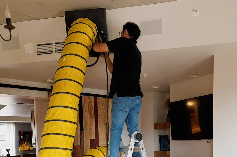 A technician on a ladder running a yellow vacuum hose into a ceiling duct opening inside a home.