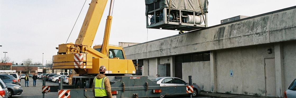 A mobile crane on outriggers lifting a packaged rooftop unit onto a commercial roof.