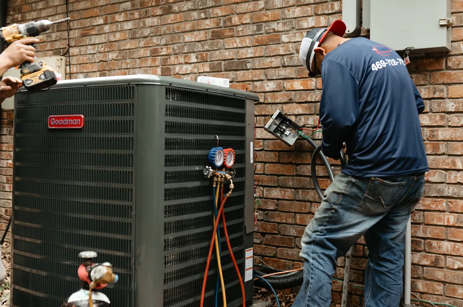 A technician connecting manifold gauges to an outdoor condenser beside a brick home while a second tech drills overhead.