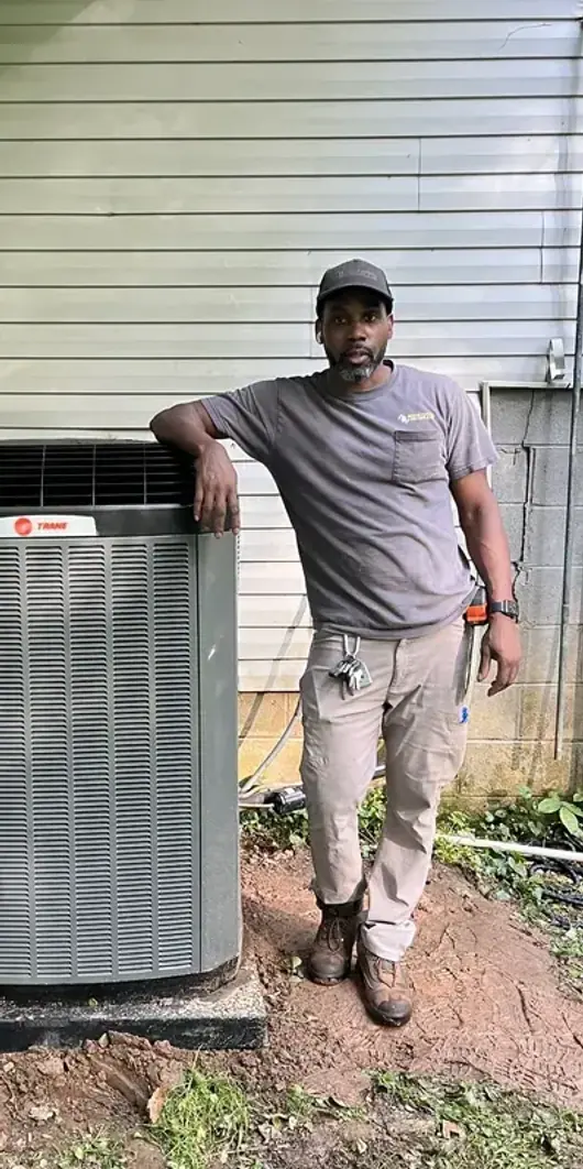 An HVAC technician leaning on an outdoor condenser unit he has just installed beside a house