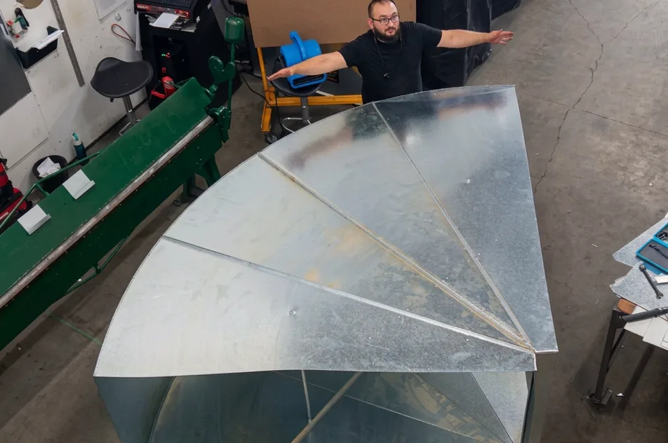 A fabricator in a workshop standing behind a large galvanized sheet metal radius fitting.