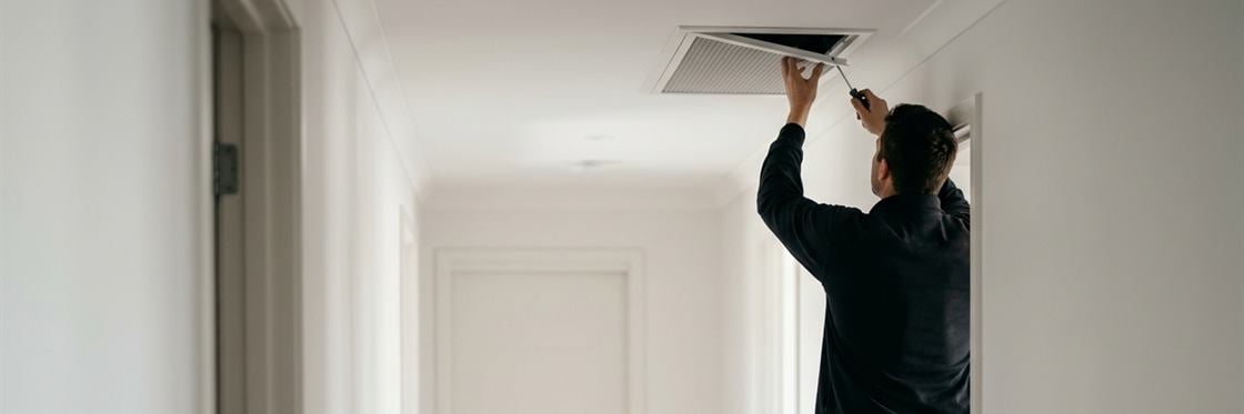 A technician on a step ladder removing a ceiling supply air register in a clean hallway.