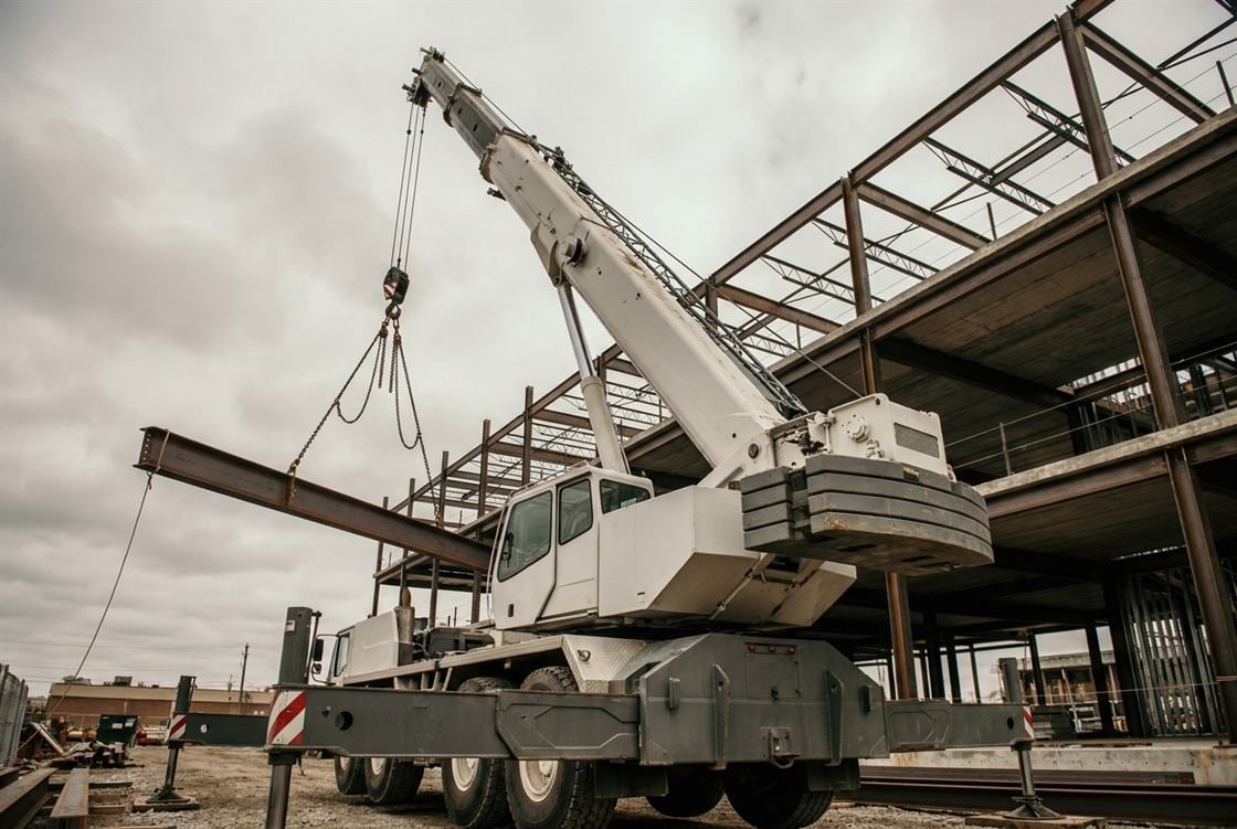 A mobile hydraulic crane on outriggers at a construction site with its boom extended, lifting a steel load while the rigging crew guides it in